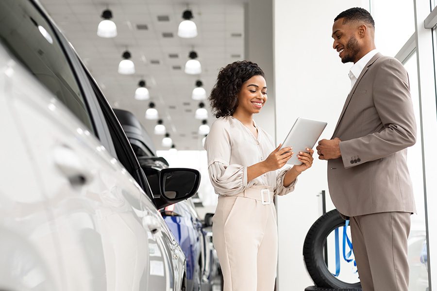 Lease Buyout Form - Professional Woman Starring at a Clipboard Smiling and a Professional Man is Smiling Next to Her in a Car Dealership on a Nice Day